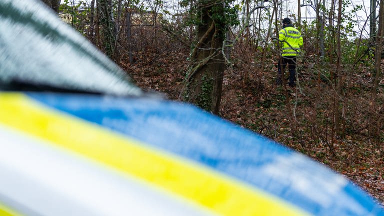 Ein Polizist steht an einem Hang im Wald, im Vordergrund unscharf zu sehen, die Motorhaube eines Polizeiautos (Symbolbild)