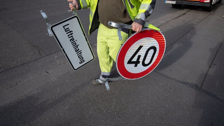 Symbolbild: Ein Mann trägt ein Tempo 40 Schild und ein Schild mit der Aufschrift "Luftreinhaltung".
