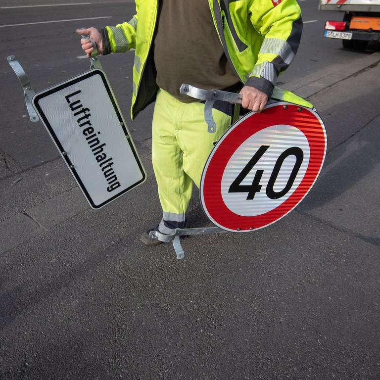 Symbolbild: Ein Mann trägt ein Tempo 40 Schild und ein Schild mit der Aufschrift "Luftreinhaltung".