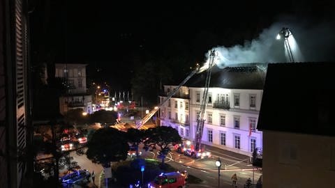 Aus dem Dachstuhl eines Hotels in Baden-Baden dringen weiße Rauchschwaden, während die Feuerwehr den Brand von Drehleitern aus bekämpft. (Foto: SWR)