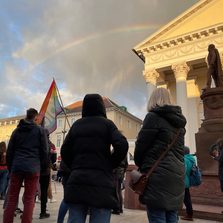 Ein Regenbogen am Himmel und Menschen halten Rregenbogenfahne in der Hand - bei der Mahnwache auf dem Karlsruher Marktplatz stehen Menschen bei einer Mahnwache gegen den Krieg in der Ukraine (Foto: SWR, SWR)
