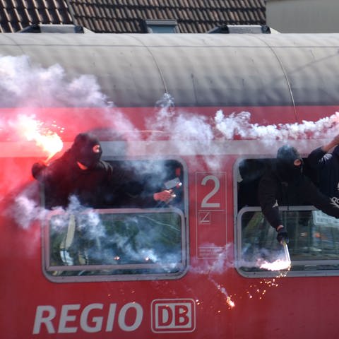 Drei vermummte Ultra-Fans in einem roten DB-Regionalzug, die Bengalo-Feuer entzündet haben und aus dem Fenster gucken. (Foto: picture-alliance / Reportdienste, picture alliance / Andreas Rosar/dpa | Andreas Rosar)