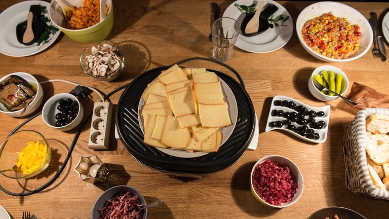 Raclettekäse und Schalen mit Zutaten stehen auf einem Tisch. (Foto: dpa Bildfunk, picture alliance / Patrick Seeger/dpa | Patrick Seeger)