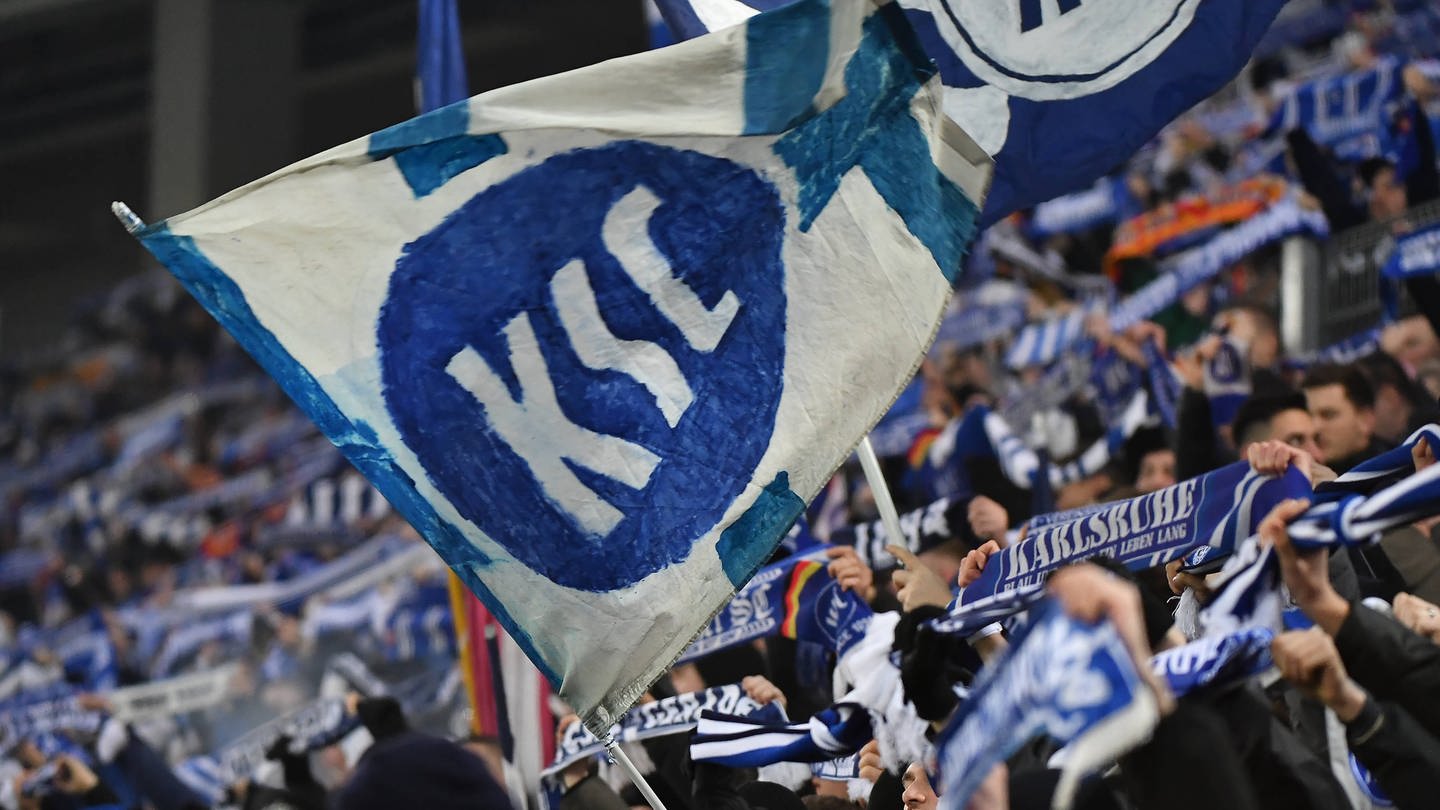 Eine KSC-Fahne im Vordergrund, im Hintergund unscharf KSC-Fans mit Schals auf der Tribüne. (Foto: IMAGO, Zink)