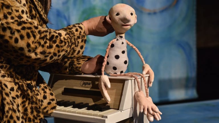 Eine stilisierte Puppe mit dünnen Ärmchen und Beinchen und einenm birnenförmigen Körper mit schwarzen Punkten, sitzt af einem Miniatur-Klavier. (Foto: Pressestelle, Theater Flunker-Produktionen)
