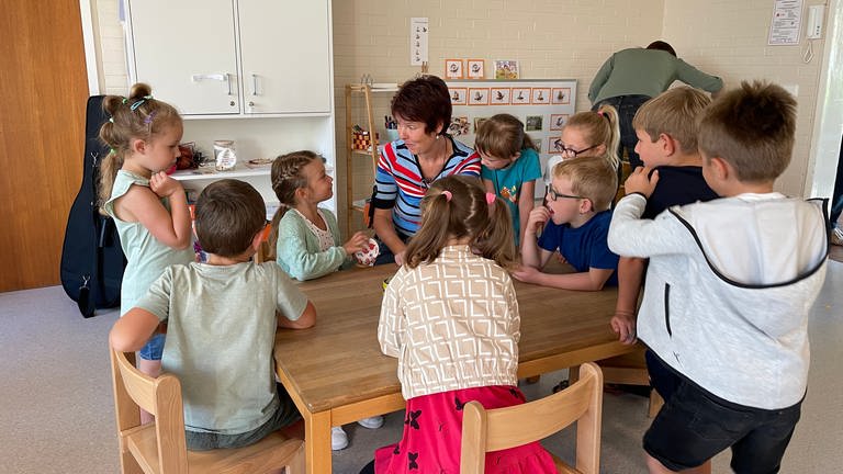 Viele Kinder sitzen rund um einen Tisch zusammen mit einer Erzieherin (Foto: SWR, Tabea Spengler)