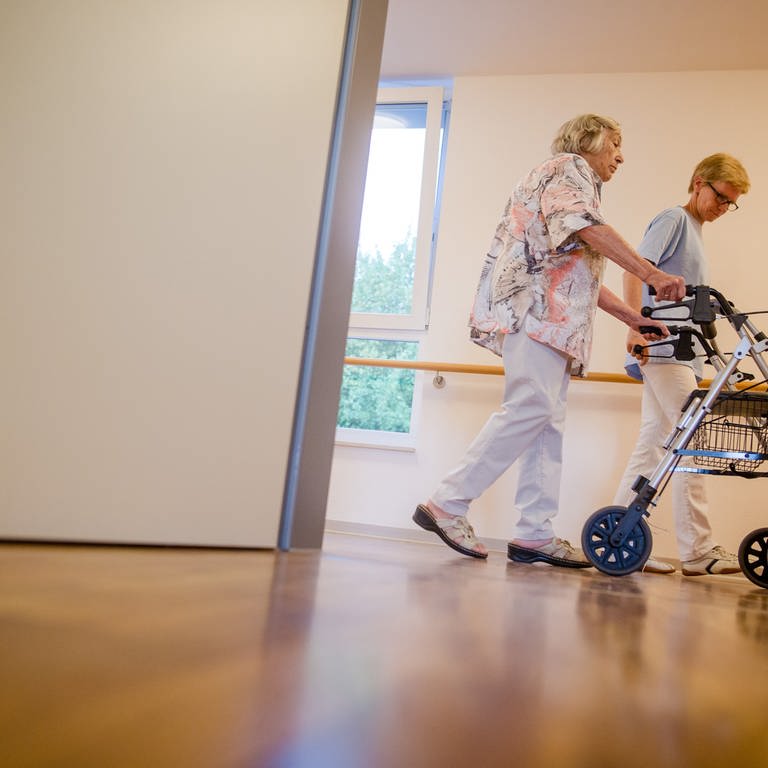 Eine Pflegekraft geht in einem Pflegeheim mit einer älteren Dame über einen Korridor. (Foto: picture-alliance / Reportdienste, SWR, picture alliance/dpa | Christoph Schmidt)