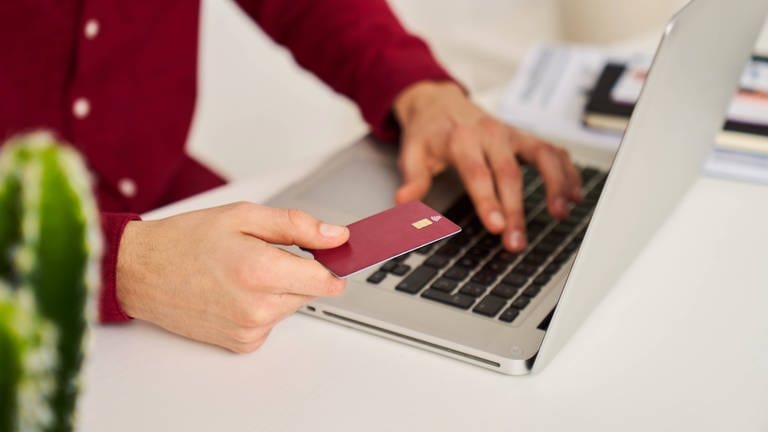 Ein Mann hält eine Kreditkarte in der Hand und hat einen aufgeklappten Laptop neben sich. (Foto: IMAGO, IMAGO / Addictive Stock)