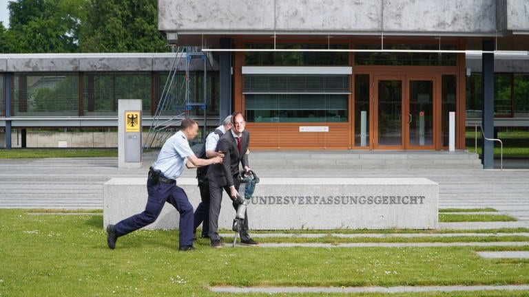 Ein als Bundeskanzler Olaf Scholz verkleideter Klimaaktivist der Gruppierung "Letzte Generation" wird am Montag bei einer Aktion vor dem Bundesverfassungsgericht in Karlsruhe von Sicherheitskräften abgeführt. 