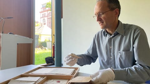 Martin Stingl vom Generallandesarchiv in Karlsruhe überprüft an seinem Schreibtisch die gefundenen Dokumente auf ihre Echtheit. Dabei trägt er weiße Stoffhabdschuhe. (Foto: SWR)