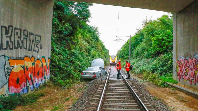 Ein Auto liegt unter einer Brücke auf den Gleisen. Vor dem Fahrzeug befindet sich ein Zug auf den Gleisen. (Foto: Pressestelle, Waldemar Gress / EinsatzReport24)