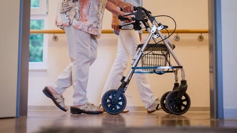 Eine Frau mit Rollator läuft zusammen mit einem Altenpfleger den Gang entlang. Auch in der Pflege in Karlsruhe ist Zeitarbeit mittlerweile Teil des Alltags. (Foto: dpa Bildfunk, Picture Alliance / Christoph Schmidt)
