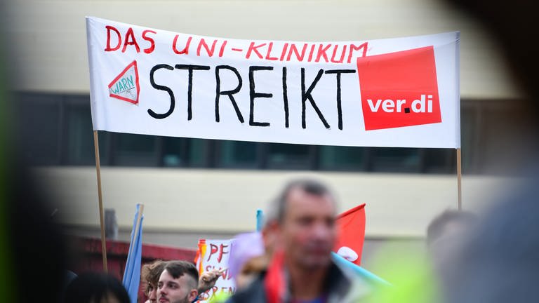 Streikende Beschäftigte halten bei einer Kundgebung vor dem Universitätsklinikum Heidelberg ein Transparent mit der Aufschrift «Das Uni-Klinikum streikt» in die Höhe (Foto: dpa Bildfunk, picture alliance/dpa | Uwe Anspach)