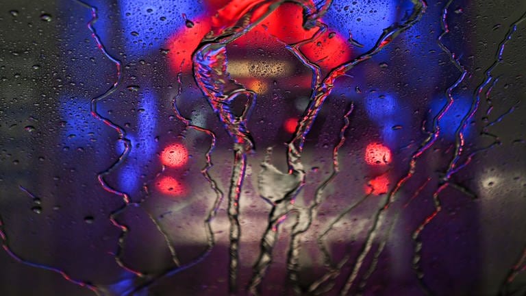 Wasser perlt an der Frontscheibe eines Pkw ab, der durch eine in rotes und blaues Licht getauchte Waschanlage fährt.