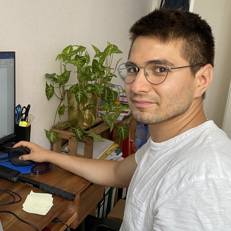 Karim Baghlani am Schreibtisch vor Computer mit Maus in der Hand und Pflanze im Hintergrund