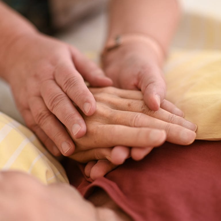 Eine Hospizmitarbeiterin hält die Hand eines todkranken Menschen, der in einem Hospiz im Bett liegt (Foto: dpa Bildfunk, picture alliance/dpa | Felix Kästle)