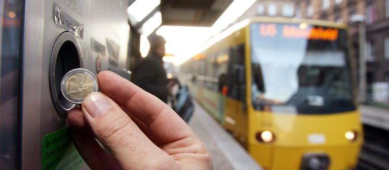 Eine Hand, die eine zwei Euro Münze an einen Fahrkartenautomaten der Straßenbahn einwirft. Im Hintergrund fährt eine Stadtbahn. (Foto: dpa Bildfunk, picture alliance/Bernd Weissbrod/dpa)