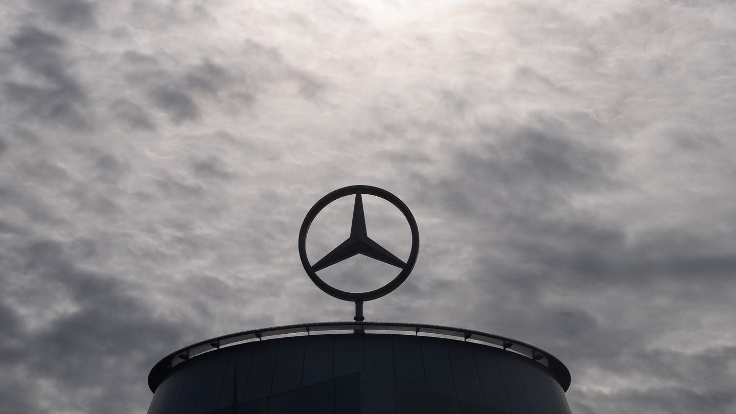Ein Mercedes-Stern ist auf der Mercedes-Benz Hauptniederlassung in Stuttgart vor dunklen Wolken und Abluft, die aus einem Industrieschornstein kommt, zu sehen. (Foto: dpa Bildfunk, picture alliance/dpa | Marijan Murat)