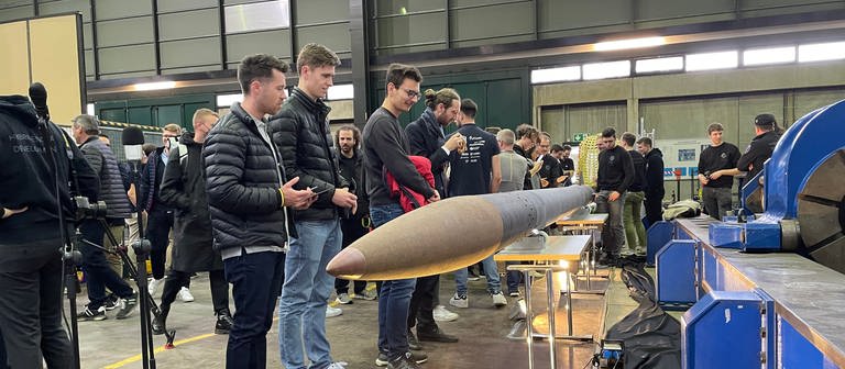 Studierende stehen seitlich neben der quer, liegenden Rakete und betrachten sie.  (Foto: SWR)