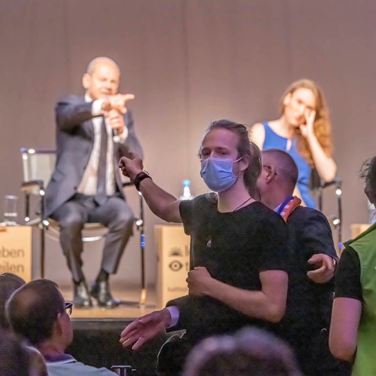 Mann zeigt im Publikum auf Kanzler Scholz auf der Bühne (Foto: IMAGO, IMAGO / Arnulf Hettrich)