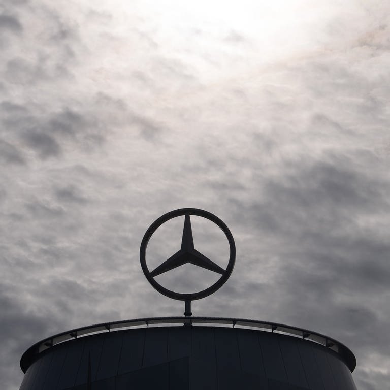 Ein Mercedes-Stern ist auf der Mercedes-Benz Hauptniederlassung in Stuttgart vor dunklen Wolken und Abluft, die aus einem Industrieschornstein kommt, zu sehen. (Foto: dpa Bildfunk, picture alliance/dpa | Marijan Murat)