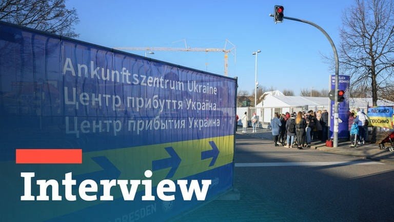 Geflüchtete Menschen aus der Ukraine warten vor einem Ankunftszentrum auf den Einlass, auf den große Schilder in blau und gelb, den ukrainischen Nationalfarben, und in dreisprachigen Schriftzügen hinweisen.