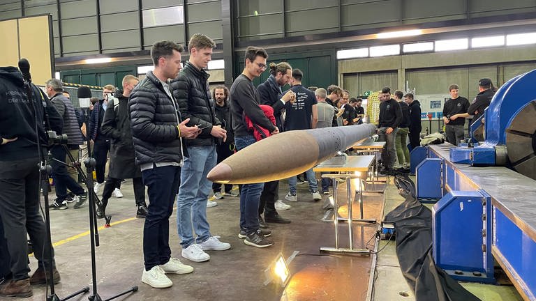 Studierende stehen seitlich neben der quer, liegenden Rakete und betrachten sie.  (Foto: SWR)