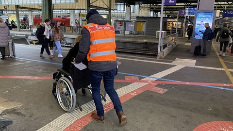 Ein Service-Mitarbeiter der Deutschen Bahn schiebt eine Rollstuhlfahrerin zu ihrem Gleis. 