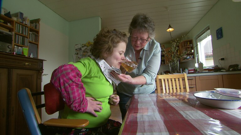 Ursula Hofmann kümmert sich seit 21 Jahren im ihre schwerbehinderte Tochter Anne. Die beiden sitzen am Esstisch und die Mutter gibt der Tochter aus einem Glas etwas zu trinken.