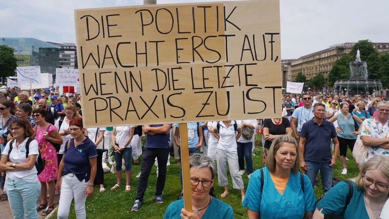 Niedergelassene Ärzte, Psychotherapeuten und Beschäftigte von Arztpraxen demonstrieren auf dem Schlossplatz in Stuttgart. Eine Teilnehmerin hält ein Schild hoch mit der Aufschrift: "Die Politik wacht erst auf, wenn die letzte Praxis zu ist." 