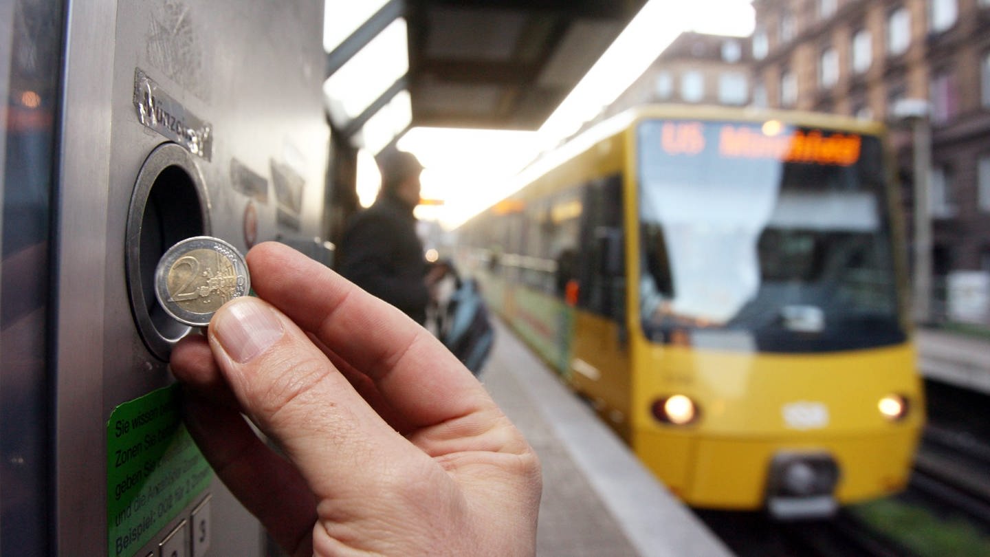 Eine Hand, die eine Münze an einen Fahrkartenautomaten der Straßenbahn einwirft. Im Hintergrund fährt eine Stadtbahn. (Foto: dpa Bildfunk, picture alliance/Bernd Weissbrod/dpa)