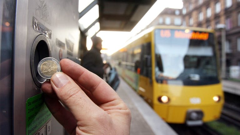 Eine Hand, die eine Münze an einen Fahrkartenautomaten der Straßenbahn einwirft. Im Hintergrund fährt eine Stadtbahn.
