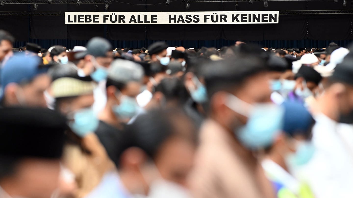 Tausende beten gemeinsam bei der Jalsa Salana 2022 in Karlsruhe. Im Hintergrund ist ein Banner mit der Aufschrift 