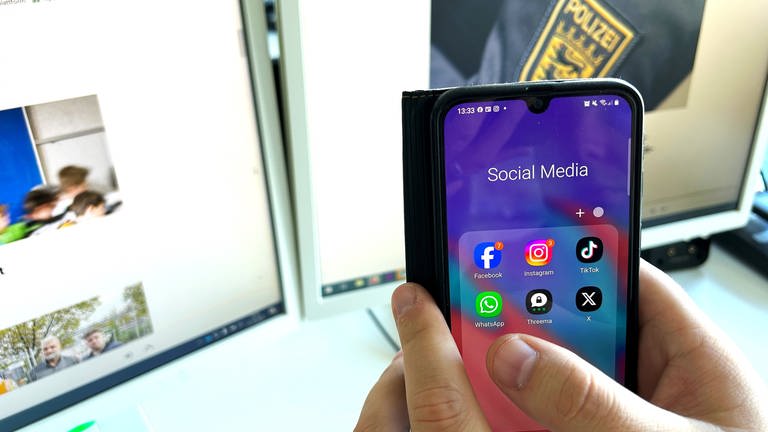 Symbolbild Faktencheck: Eine Hand mit Handy mit Social Media Apps vor einem Bildschirm. 