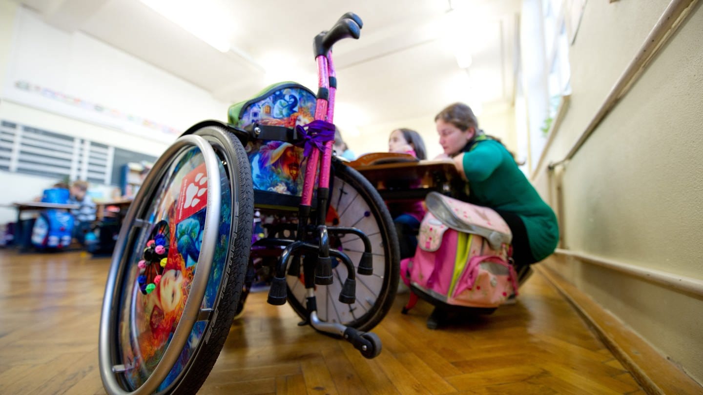 Eine junge Frau, die ein Freiwilliges Soziales Jahr (FSJ) macht, betreut in einem Klassenzimmer einer Gemeinschaftsschule in Stuttgart eine Grundschülerin, die auf einen Rollstuhl angewiesen ist (Archivbild). (Foto: dpa Bildfunk, picture alliance / dpa | Inga Kjer)