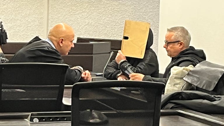 Der Angeklagte hält einen beige-farbenen Ordner vor sein Gesicht - beim Prozessauftakt im Landgericht Stuttgart.