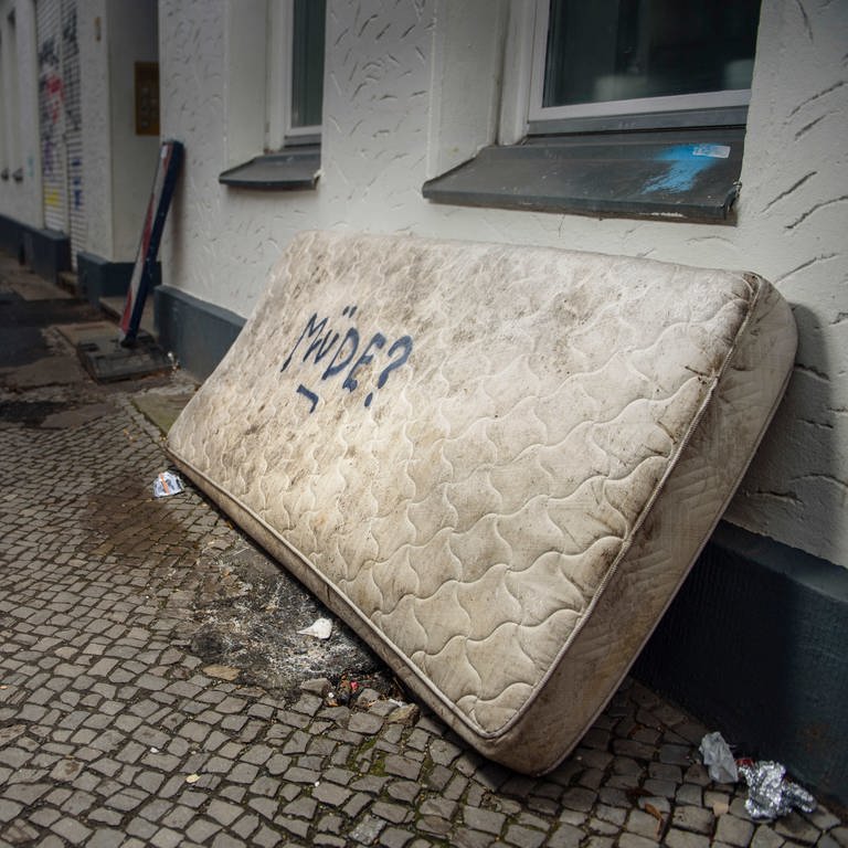 eine ausrangierte Matratze mit der Aufschrift "Müde?" auf einem Gehweg einer Großstadt (Archivbild) (Foto: dpa Bildfunk, picture alliance/dpa | Gregor Fischer (Archivbild))