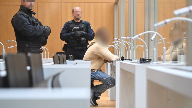 Der Angeklagte im Gerichtssaal in Stuttgart-Stammheim zu Beginn seines Prozesses. Neben ihm stehen zwei Justizbeamte. Es ist das erste Verfahren, mit dem die Eritrea-Ausschreitungen in Stuttgart im Sommer 2023 aufgearbeitet werden.