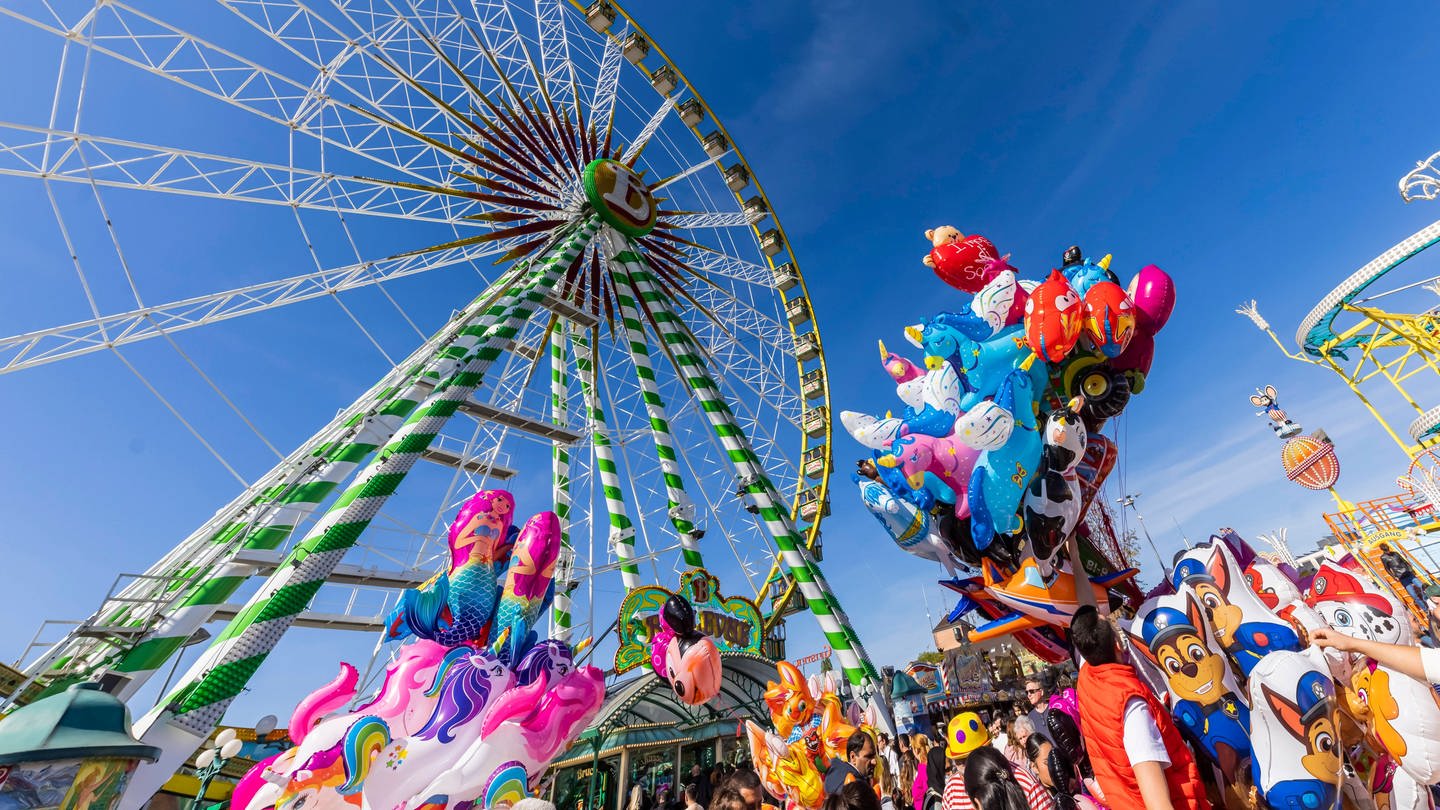 Das Datum vom Frühlingsfest 2024 in Stuttgart auf dem Wasen: 20. April bis 12. Mai. Wer ins Zelt will, sollte einen Tisch reservieren. Der Bierpreis ist gestiegen. Zu sehen sind auf dem Bild das Riesenrad, Luftballons, Menschen und die Wilde Maus.