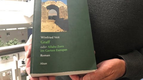 Das grüne Buch mit dem Titel: "Graff: oder Allahs Zorn im Garten Europas" wird von zwei Händen gehalten, vor einem schwarzen Pullover. (Foto: SWR, Dinah Steinbrink)