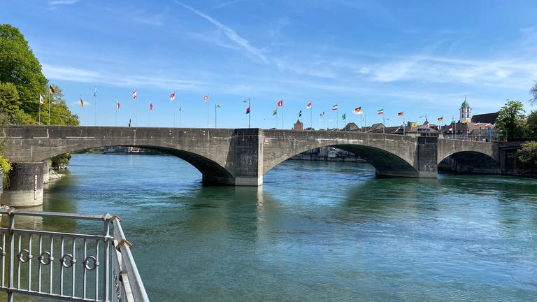 Zu sehen ist die steinerne Rheinbrücke, sie verbindet das Schweizer und deutsche Rheinfelden. Auf der Brücke wehen bunte Fahnen. Der Rhein schimmert blau und grün, der Himmel ist fast wolkenlos. (Foto: SWR, Katharina Seeburger)