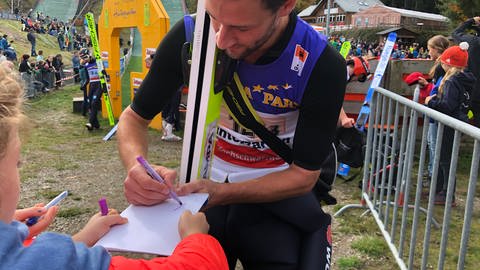 Markus Eisenblichler, der noch seine Skier im Arm hält, unterschreibt auf einem Block, den ihm ein Kind entgegenstreckt. (Foto: SWR, Dinah Steinbrink)