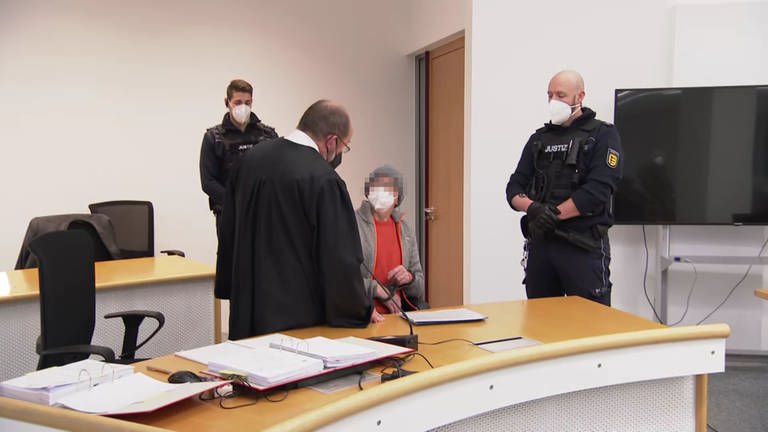 Der mutmaßlicher "Reichsbürger" sitzt zu Prozessbeginn im Gerichtssaal des Oberlandesgerichts in Stuttgart auf einem Stuhl. Er trägt einen orangenen Pullover, darüber eine graue Jacke. Neben ihm steht sein Verteidiger, der trägt die schwarze Anwaltsrobe. Neben den beiden stehen zwei Polizisten. Alle tragen FFP2-Masken. (Foto: SWR)