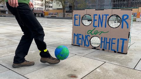 Ein Mann schießt einen Fußball, der wie eine Weltkugel aussieht. Auf dem Tor steht "Eigentor der Menschheit. (Foto: SWR, Paula Zeiler)