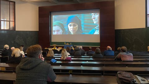 In einem Hörsaal der Freiburger Universität sitzen Menschen, auf den Tischen stehen kleine blau-gelbe Fahnen der Ukraine. Die Menschen sind von hinten zu sehen und blicken auf eine Leinwand. Auf der Leinwand wird ein Film über den Holodomor gezeigt. In der Szene sind zwei verzweifelte Frauen zu sehen. (Foto: SWR, Sebastian Bargon)