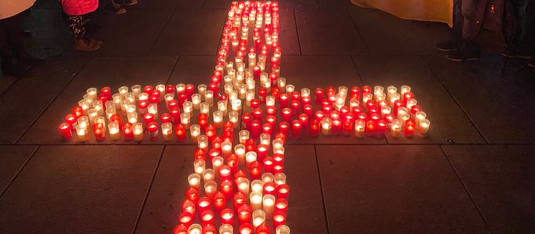In der Mitte des Bildes, auf dem Boden, stehen weiße und rote Kerzen. Sie sind zu einem Kreuz angeordnet. Um das Kreuz herum stehen Erwachsene und Kinder, sie halten die blau-gelben Fahnen der Ukraine in der Hand. Unter ihnen ist auch ein Pfarrer in Talar. (Foto: SWR, Sebastian Bargon)