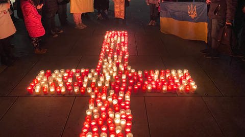In der Mitte des Bildes, auf dem Boden, stehen weiße und rote Kerzen. Sie sind zu einem Kreuz angeordnet. Um das Kreuz herum stehen Erwachsene und Kinder, sie halten die blau-gelben Fahnen der Ukraine in der Hand. Unter ihnen ist auch ein Pfarrer in Talar. (Foto: SWR, Sebastian Bargon)