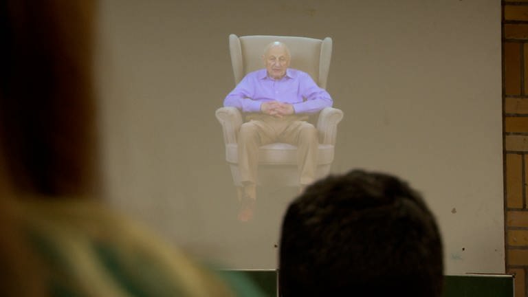 Ein älterer Mann in einem lila Hemd sitzt auf einem großen Sessel. (Foto: SWR)