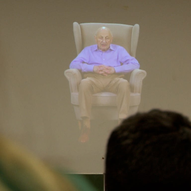 Ein älterer Mann in einem lila Hemd sitzt auf einem großen Sessel. (Foto: SWR)