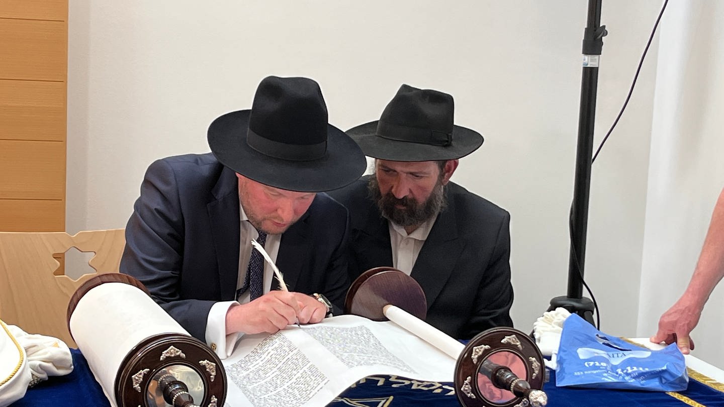 Die Torarolle liegt auf einem Tisch in der Rottweiler Synagoge während Landesrabbiner Moshe Flomenmann mit Unterstützung von Sofer Boruch Lamdan die letzten 12 Buchstaben mit einer Feder auf das Pergament geschrieben werden. (Foto: SWR, Hardy Faißt)